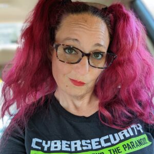 Headshot of Bambi Okugawa, a smiling woman with bright magenta hair styled in pigtails, wearing glasses and a black cybersecurity t-shirt.